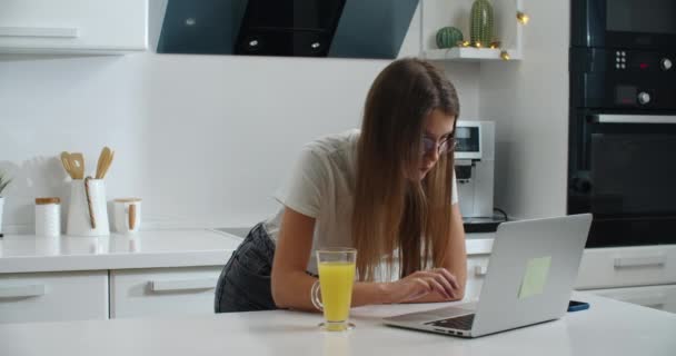 Een jonge vrouw met een bril staat bij de tafel thuis en werkt op een laptop. Maakt het werk af en sluit de laptop. Drinkt sap en lacht. Werk vanuit huis — Stockvideo