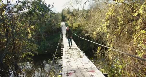 Un touriste mâle marche le long d'un pont suspendu en bois lors d'une soirée d'automne. — Video