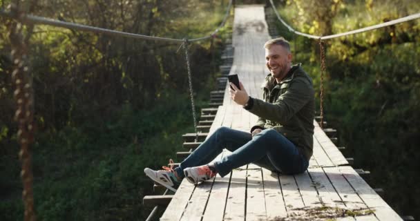 Samiec turysty siedzi na drewnianym moście wiszącym i komunikuje się z przyjaciółmi przez komunikację wideo. Na tle jesiennego lasu — Wideo stockowe