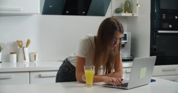 Une jeune femme avec des lunettes se tient près de la table à la maison et travaille sur un ordinateur portable. Termine le travail et ferme l'ordinateur portable. Il boit du jus. Travail à domicile — Video