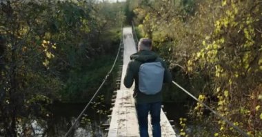 Sırt çantalı bir erkek turist, bir sonbahar akşamı asılmış ahşap bir köprü boyunca yürüyor. Arka plan