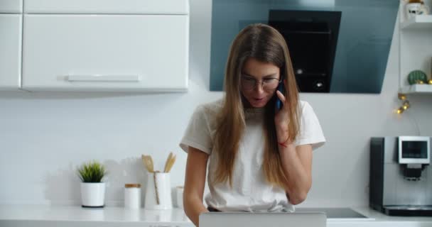 Een jonge studente die thuis aan de laptop werkt en aan de telefoon praat. Werk vanuit huis. — Stockvideo