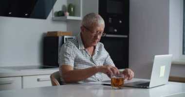 Gözlüklü yaşlı adam internetten alışveriş yapmak ve kredi kartıyla ödeme yapmak için dizüstü bilgisayar kullanıyor. Ev mutfağı.