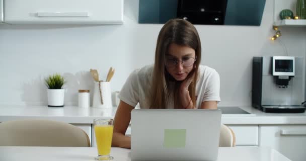 Μια νεαρή γυναίκα με γυαλιά στέκεται στο τραπέζι στο σπίτι και εργάζεται σε ένα φορητό υπολογιστή. Μερικές φορές πίνει χυμό. Εργασία από το σπίτι — Αρχείο Βίντεο