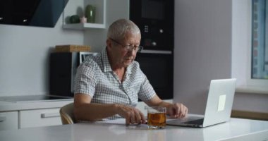 Gözlüklü yaşlı adam internetten alışveriş yapmak ve kredi kartıyla ödeme yapmak için dizüstü bilgisayar kullanıyor. Ev mutfağı.