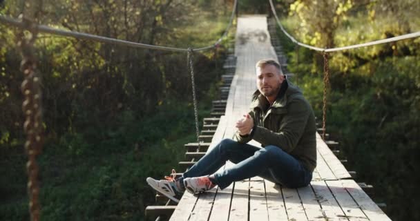 一个男人穿着一件暖和的夹克坐在一座木头桥上，暖手。在绿色植物的背景下. — 图库视频影像