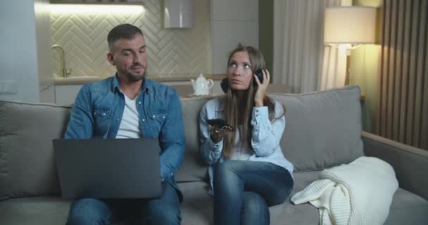 A férj és a feleség együtt töltik az estét otthon. A férj egy laptopon dolgozik, a feleség pedig fejhallgatón hallgat zenét. Családi idő. — Stock videók