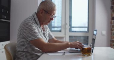 Evde dizüstü bilgisayarla çalışan gözlüklü yaşlı iş adamı portresi. Bir adam dizüstü bilgisayarda çalışır, notlar alır ve meyve suyu içer. Evden çalış..