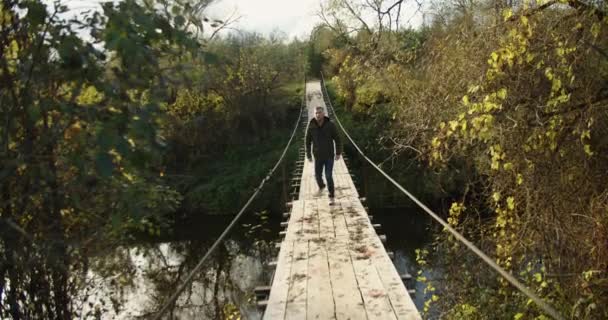 一个穿着保暖夹克的徒步旅行者在一座木桥上行走。在绿色植物的背景下. — 图库视频影像