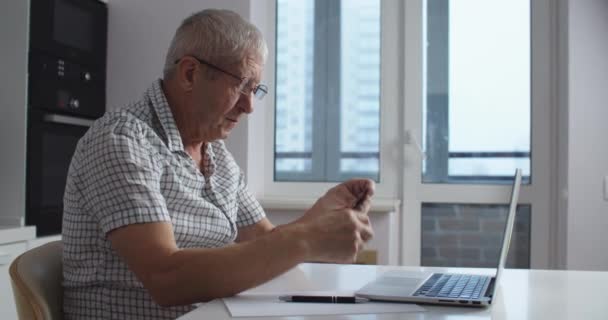 Hombre mayor con gafas usando ordenador portátil hace compras en línea y pagar con tarjeta de crédito. Cocina casera. — Vídeos de Stock