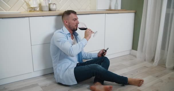 Seorang pria duduk di lantai dapur, minum anggur merah dan membaca pesan di ponselnya. Kesenangan di rumah saja. — Stok Video