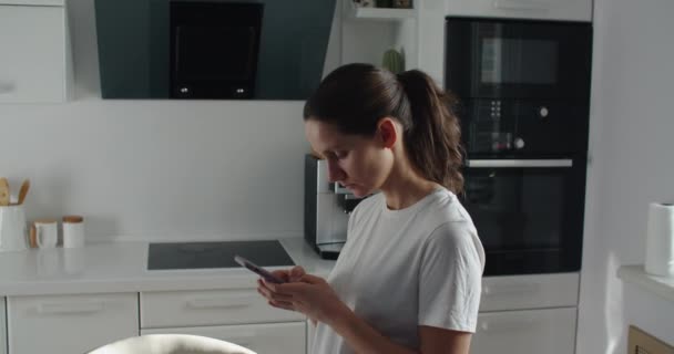 Een vermoeide vrouw staat in de keuken en schrijft een bericht op haar mobiele telefoon. — Stockvideo