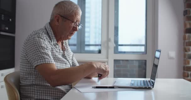 Senior man met een bril met laptop computer doet online winkelen en betalen met een creditcard. Huiskeuken. — Stockvideo