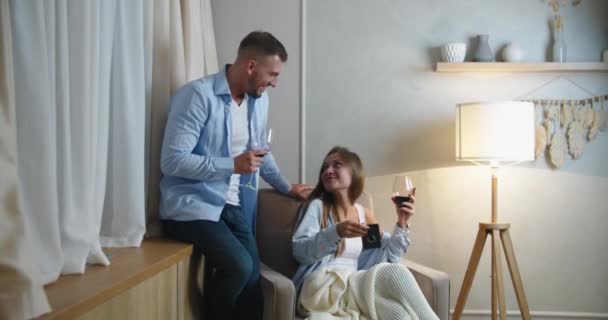 Lächelnd sitzen junge Eheleute in einem Stuhl, trinken Rotwein und genießen die Familienfreizeit am heimischen Wochenende, glückliches Millennial-Paar feiert Hochzeitstag oder romantisches Date zu Hause — Stockvideo