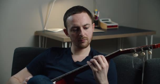 Ein junger Mann sitzt auf dem Sofa und spielt Gitarre. Nahaufnahme Porträt eines Mannes mit einer Gitarre in der Hand. — Stockvideo