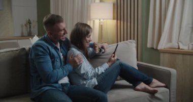 Kanepede oturan sevimli, neşeli, pozitif stil sahibi eşlerin yakın plan portresi. Kadın, erkeğe cihaz gösteriyor ve modern ışık dairelerini daktilo ediyor.