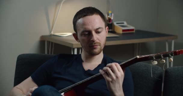 En ung man sitter på soffan och spelar gitarr. Närbild porträtt av en man med en gitarr i händerna. — Stockvideo