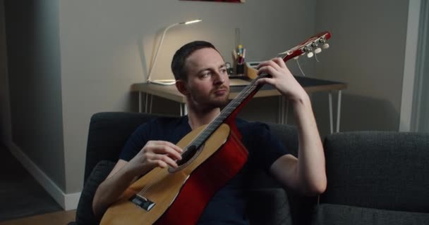 Um jovem senta-se no sofá e toca guitarra. Close-up retrato de um homem com uma guitarra em suas mãos. — Vídeo de Stock