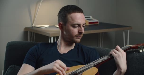Seorang pemuda duduk di sofa dan bermain gitar. Potret close-up seorang pria dengan gitar di tangannya. — Stok Video