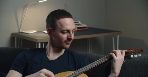 Seorang pemuda duduk di sofa dan bermain gitar. Potret close-up seorang pria dengan gitar di tangannya. — Stok Video