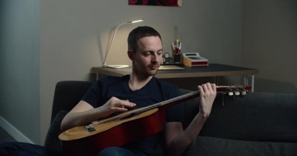 En ung man sitter på soffan och spelar gitarr. Närbild porträtt av en man med en gitarr i händerna. — Stockvideo