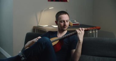 Genç bir adam kanepede oturur ve gitar çalar. Elinde gitar olan bir adamın yakın plan portresi..