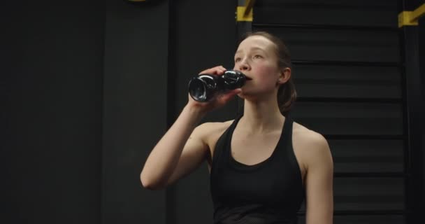 Durstige schlanke athletische Frau trinkt nach dem aktiven Training Wasser, hält eine schwarze Flasche in Sportbekleidung isoliert über eine graue Betonwand. Konzept Mensch und Müdigkeit. — Stockvideo