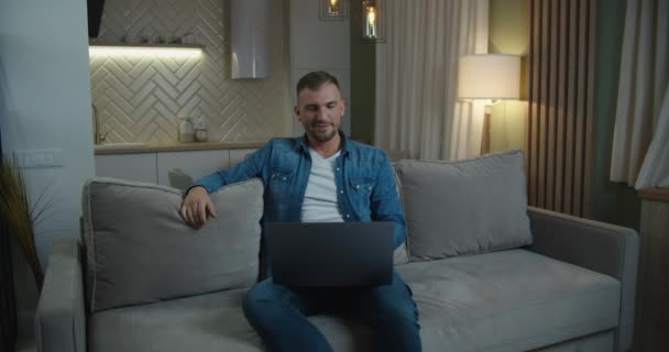 白いシャツを着たハッピーマンはソファに座って、自宅でラップトップを使用しています。オンライン作業。オンライン学習. — ストック動画