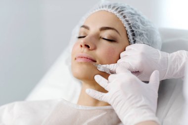 Çekici genç bir kadın canlandırıcı yüz enjeksiyonu yaptırıyor. O, klinikte sakince oturuyor. Uzman güzellik uzmanı hyaluronik asitle kadın kırışıklıklarını dolduruyor..