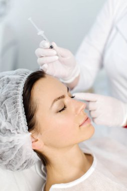 Çekici genç bir kadın canlandırıcı yüz enjeksiyonu yaptırıyor. O, klinikte sakince oturuyor. Uzman güzellik uzmanı hyaluronik asitle kadın kırışıklıklarını dolduruyor..