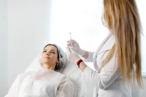 Çekici genç bir kadın canlandırıcı yüz enjeksiyonu yaptırıyor. O, klinikte sakince oturuyor. Uzman güzellik uzmanı hyaluronik asitle kadın kırışıklıklarını dolduruyor..