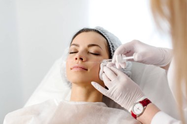 Çekici genç bir kadın canlandırıcı yüz enjeksiyonu yaptırıyor. O, klinikte sakince oturuyor. Uzman güzellik uzmanı hyaluronik asitle kadın kırışıklıklarını dolduruyor..