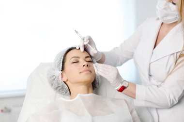 Çekici genç bir kadın canlandırıcı yüz enjeksiyonu yaptırıyor. O, klinikte sakince oturuyor. Uzman güzellik uzmanı hyaluronik asitle kadın kırışıklıklarını dolduruyor..