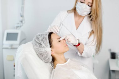 Çekici genç bir kadın canlandırıcı yüz enjeksiyonu yaptırıyor. O, klinikte sakince oturuyor. Uzman güzellik uzmanı hyaluronik asitle kadın kırışıklıklarını dolduruyor..