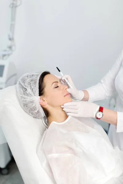 Çekici genç bir kadın canlandırıcı yüz enjeksiyonu yaptırıyor. O, klinikte sakince oturuyor. Uzman güzellik uzmanı hyaluronik asitle kadın kırışıklıklarını dolduruyor..