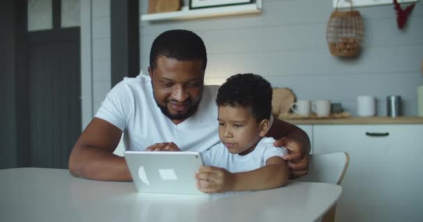 Glad afrikansk far och hans lilla son använder laptop hemma tillsammans, titta på filmer på nätet eller surfa på internet — Stockvideo