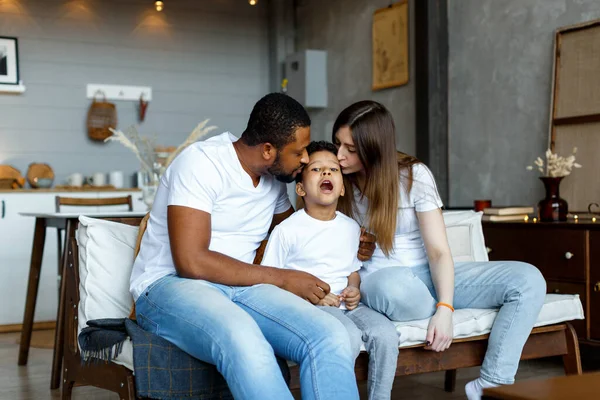 Loving black family relaxing at sofa in the morning, happy mixed race parents laughing cuddling having fun with cute little kid child son playing enjoying moments together