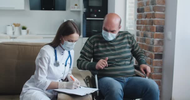 Jeune infirmière expliquant les informations à l'homme patient en fauteuil roulant tout en parlant ensemble à la maison. Concept d'épidémie et de virus — Video