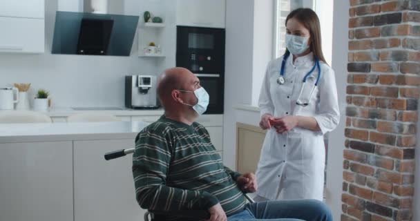Enfermeira jovem explicando informações para o homem paciente em cadeira de rodas enquanto conversam juntos em casa. Conceito de epidemia e vírus — Vídeo de Stock