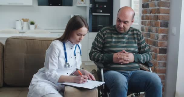 若い女性看護師が自宅で話しながら車椅子の男性患者に情報を説明する。流行とウイルスの概念 — ストック動画