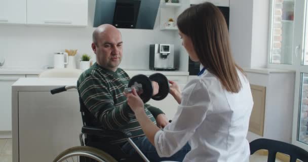 Young woman nurse explaining information to man patient in wheelchair while talking together at home. Epidemic and virus concept — Stock Video