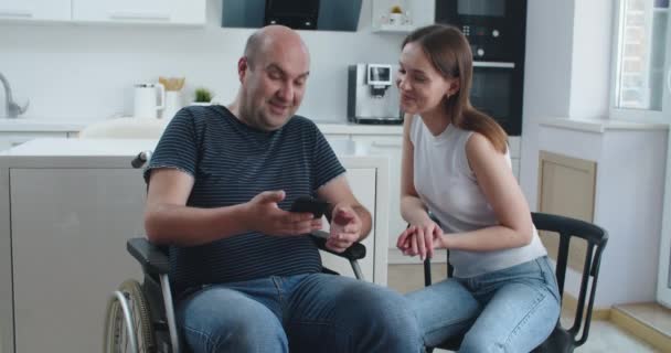Tekerlekli sandalyedeki genç adam akıllı telefondan mesaj okuyor ve kadınını gösteriyor.. — Stok video