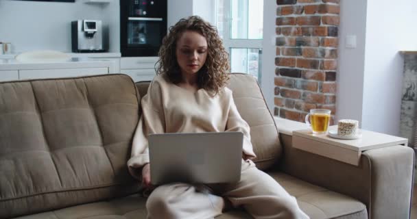 Belle fille gaie utilisant ordinateur portable en argent tout en étant assis sur le canapé dans le salon à la maison. — Video