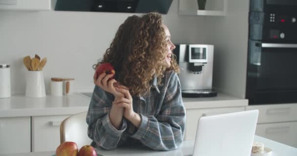 Uma garota na cozinha sorri e imprime em um laptop, depois come uma maçã. Cozinha brilhante — Vídeo de Stock