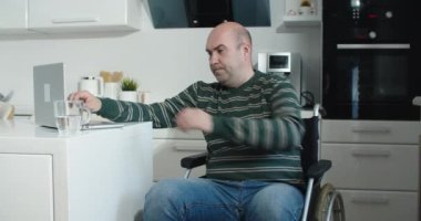 Evden tekerlekli sandalyedeki mutlu genç adam. Tekerlekli sandalyesinde oturan neşeli iş adamı. Evde dizüstü bilgisayar kullanarak tekerlekli sandalyedeki engelli adam. Masada tekerlekli sandalyede oturan genç adam..