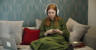 Kanepede oturup dizüstü bilgisayar ve cep telefonu kullanarak müzik dinleyen kızıl saçlı güzel bir kadın..