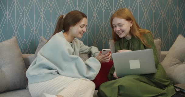 Duas jovens e alegres amigas animadas se divertindo na tela do telefone celular enquanto se sentam em um sofá confortável em casa. Amizade feminina tagarelar imagens engraçadas no celular memória agradável — Vídeo de Stock