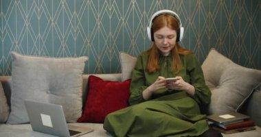 Kanepede oturup dizüstü bilgisayar ve cep telefonu kullanarak müzik dinleyen kızıl saçlı güzel bir kadın..