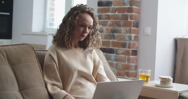 Jonge vrouw met krullend haar werkt thuis. Typen op een laptop en berichten beantwoorden op een telefoon — Stockvideo