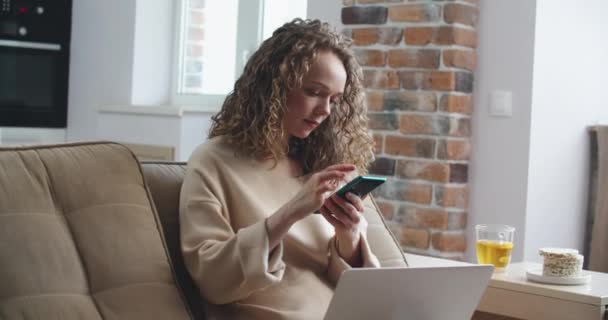 Mujer joven con el pelo rizado está trabajando en casa. Escribir en una computadora portátil y contestar mensajes en un teléfono — Vídeo de stock